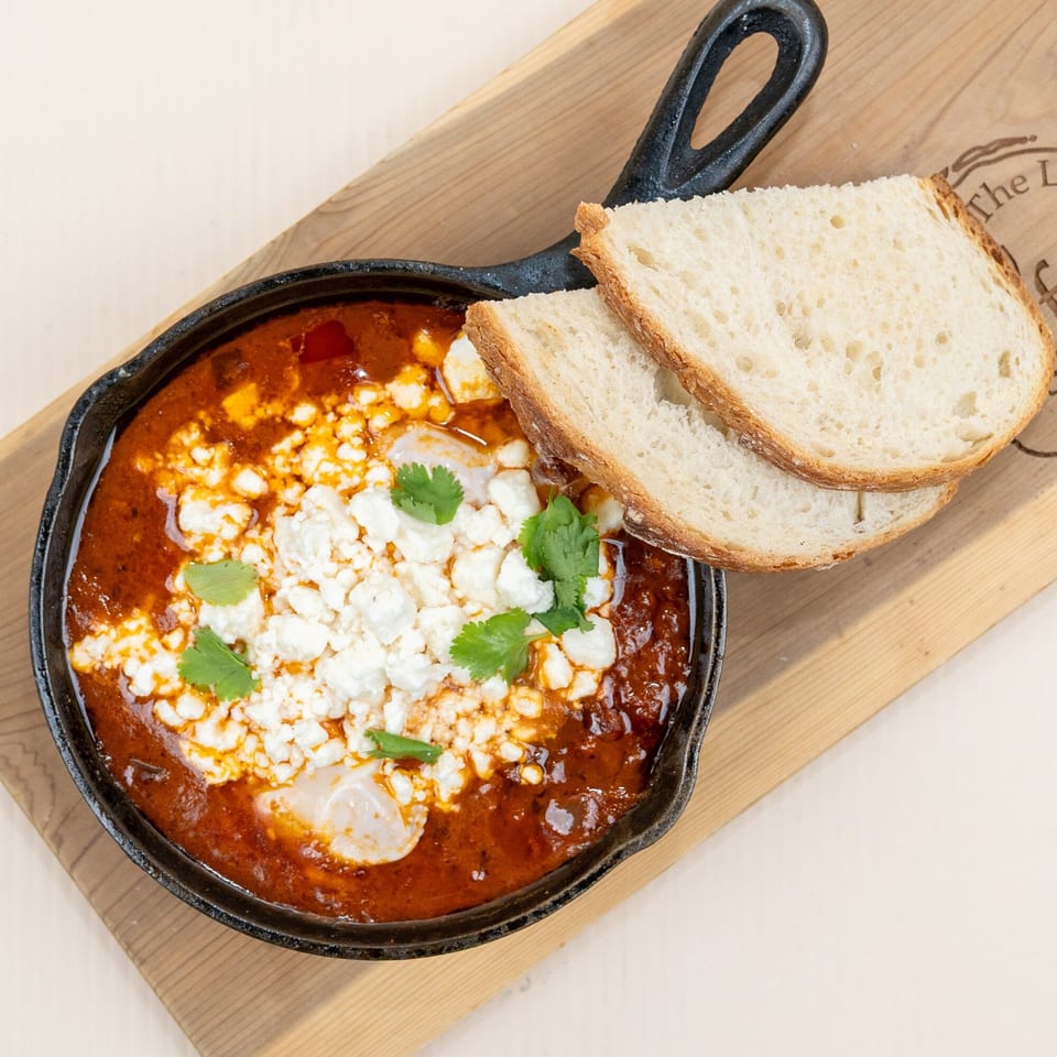 Shakshuka in a cast iron skillet with feta cheese, cilantro, and two slices of bread.