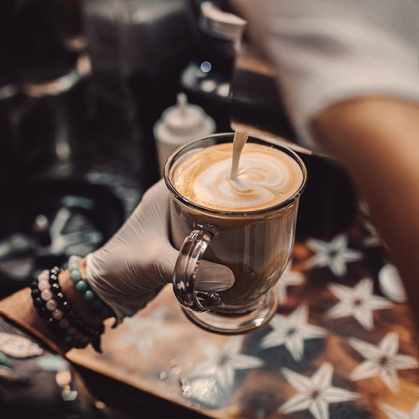A barista in a white glove pours latte art into a clear mug of coffee.
