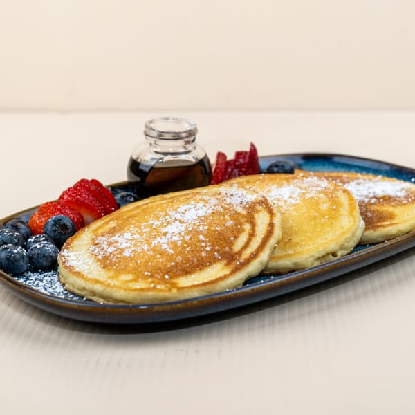 Three golden pancakes dusted with powdered sugar, served with blueberries, strawberries, and syrup.