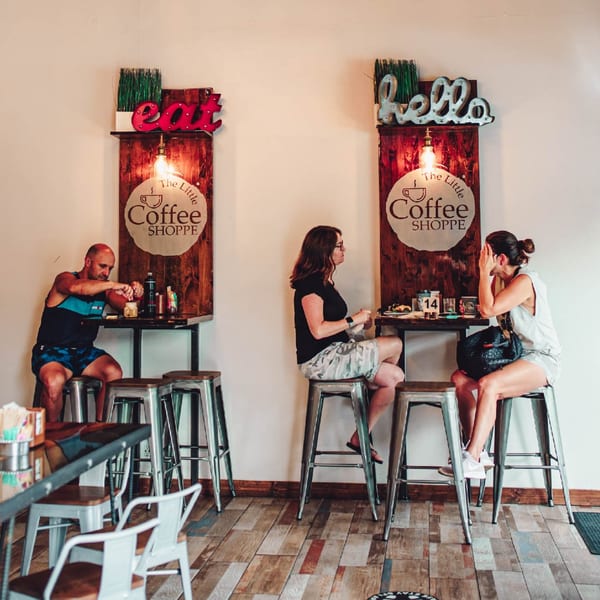 Customers sit at high-top tables with "The Little Coffee Shoppe" signs and neon "eat" and "hello" signs.