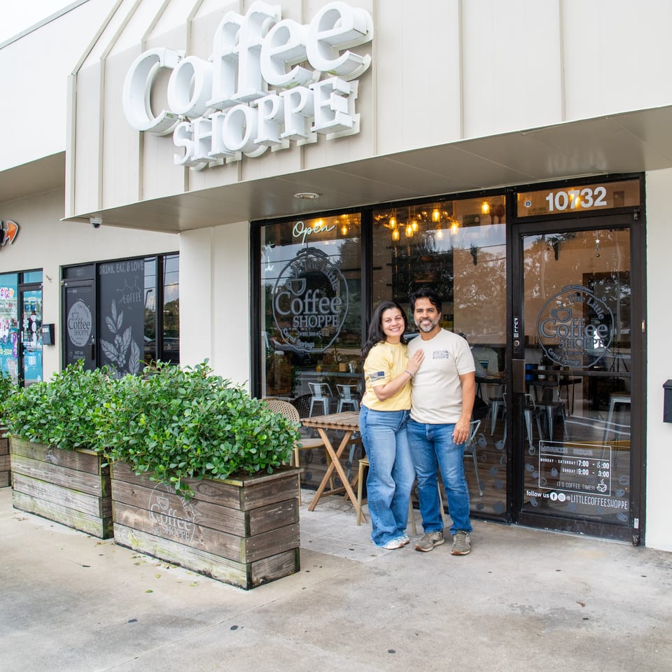 Owners of The Little Coffee Shoppe stand smiling in front of their coffee shop.