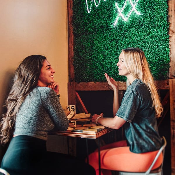Two women smiling and talking at a table in The Little Coffee Shoppe, with a neon sign on a green wall.
