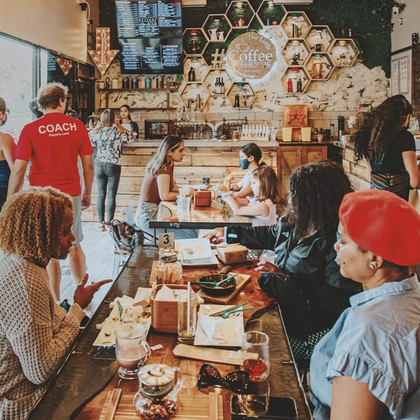 Customers enjoying coffee and food at tables inside The Little Coffee Shoppe, with a busy counter in the background.