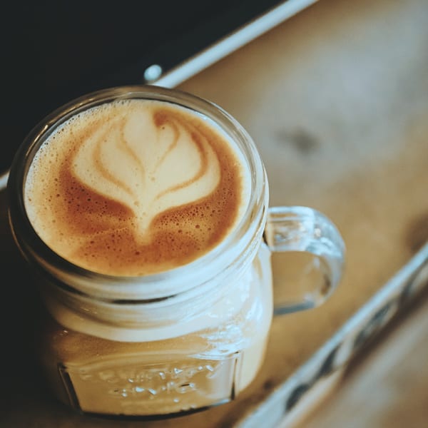 A mason jar mug of latte with heart-shaped latte art on a wooden surface.