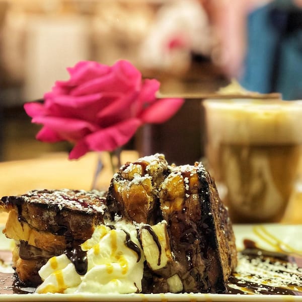 Chocolate French toast with whipped cream, caramel, and powdered sugar, with a pink rose and coffee in the background.