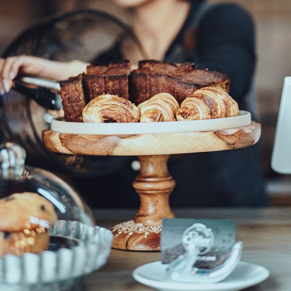 A wooden cake stand holds croissants and pastries, with a person in the background.