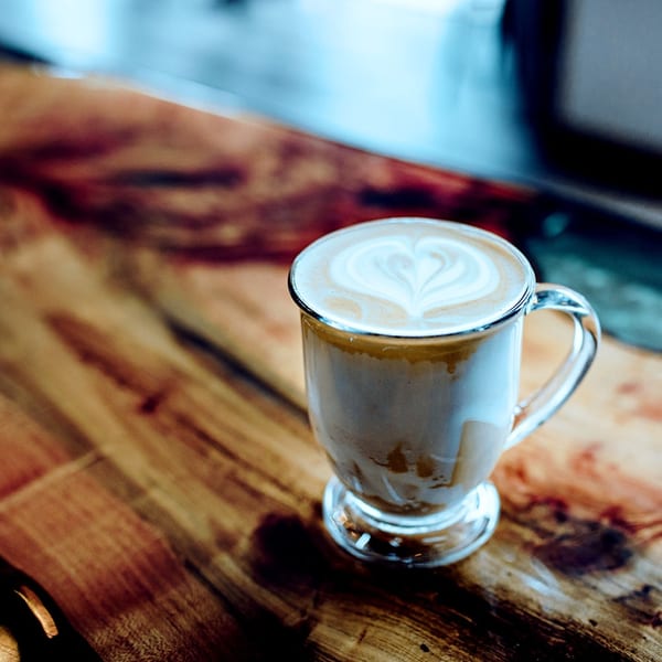 A glass mug of latte with heart-shaped latte art sits on a rustic wooden table.