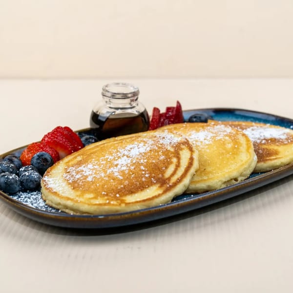 Three golden pancakes dusted with powdered sugar, fresh blueberries, strawberries, and syrup on a blue plate.