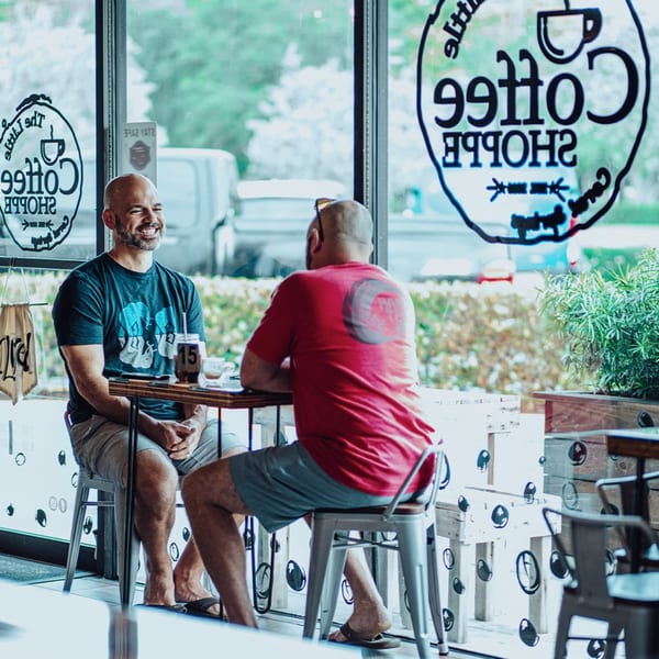 Two men sit at a table inside The Little Coffee Shoppe, one smiling at the other.