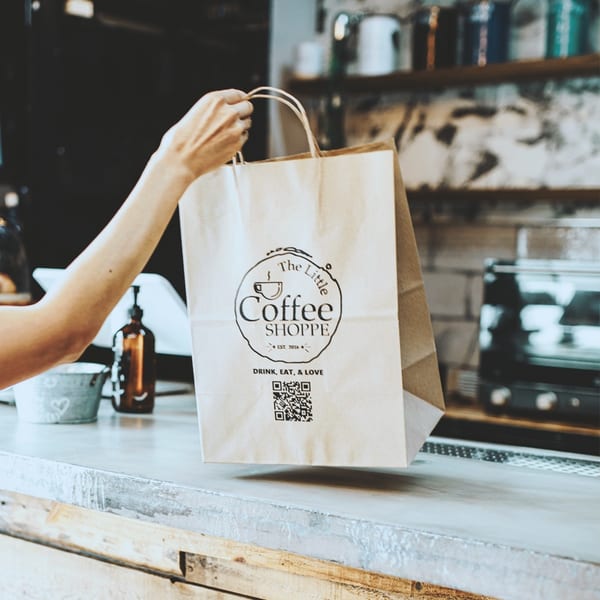 A hand holding a branded paper bag from The Little Coffee Shoppe over a counter.