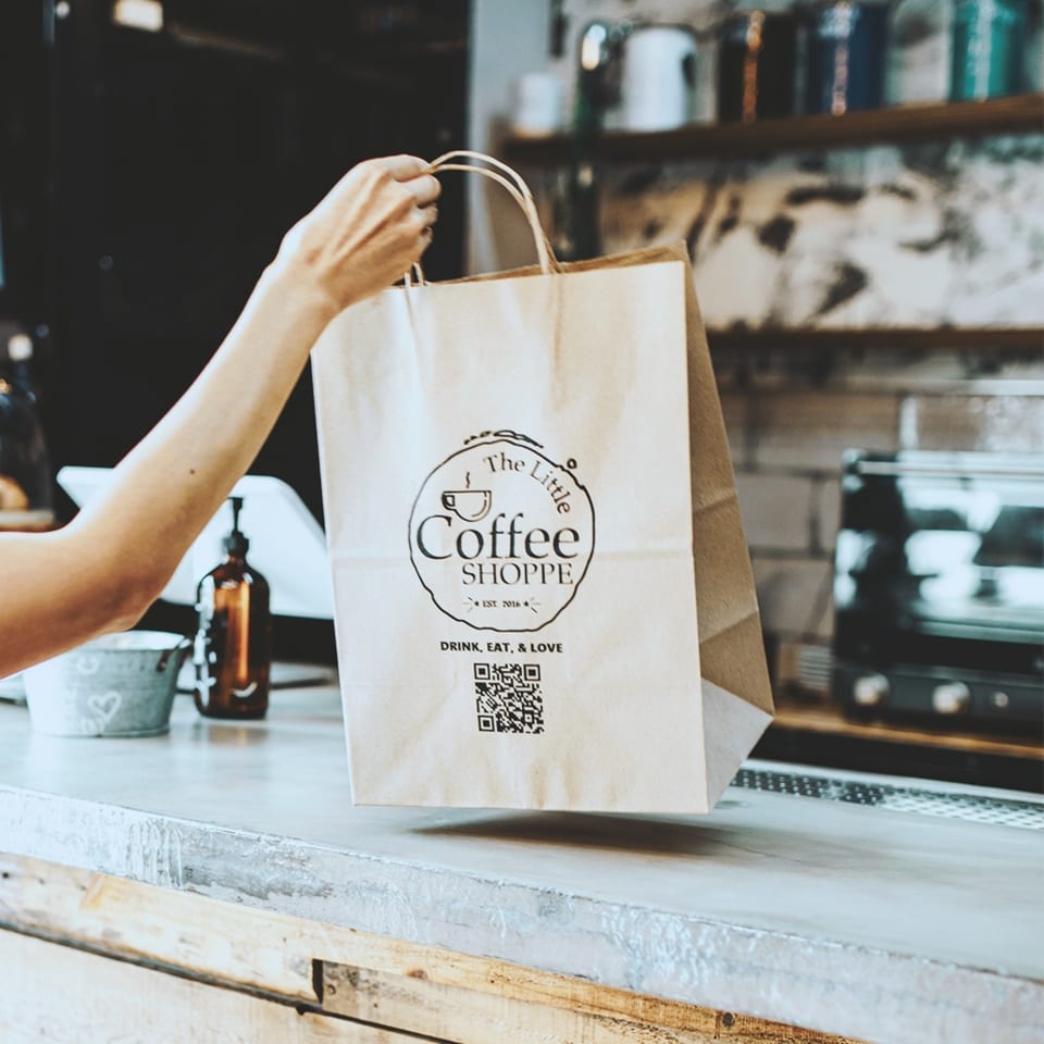 A hand holding a branded paper bag from The Little Coffee Shoppe over a counter.