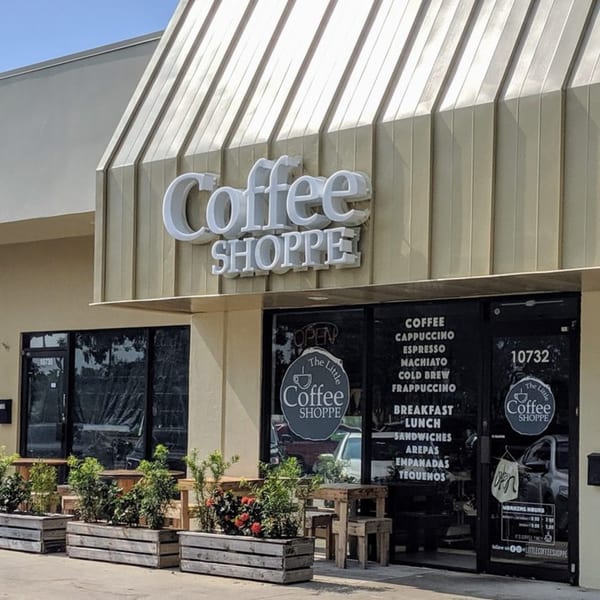 Exterior of The Little Coffee Shoppe with outdoor seating, planters, and menu items on the window.
