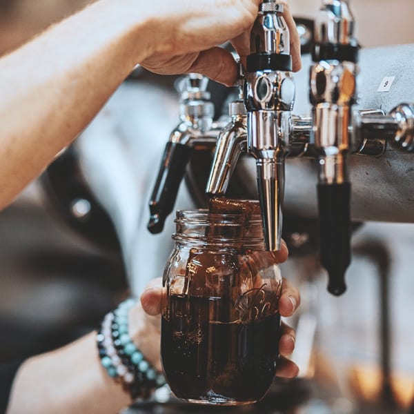 A person's hands filling a mason jar with dark coffee from a tap at The Little Coffee Shoppe.