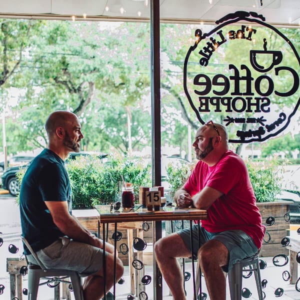 Two men sit at a table inside The Little Coffee Shoppe, seen through a window.